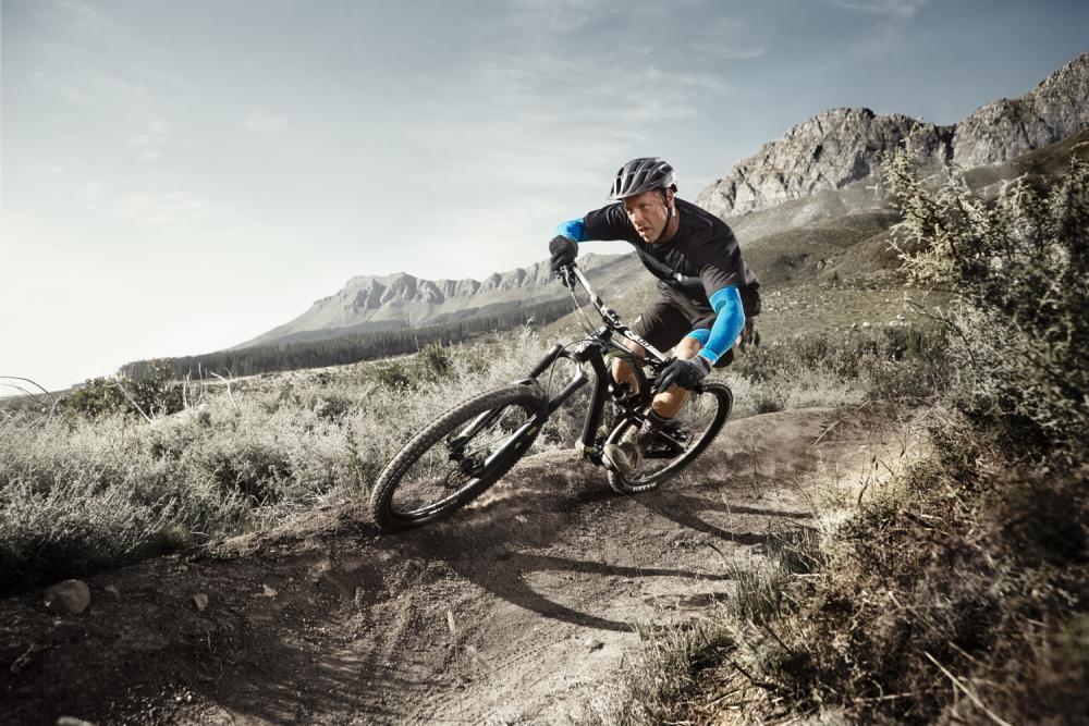 Man with Armsleeves drives on a mountain bike in an extreme corner on a dusty path through a barren landscape with bushes and mountains in the background