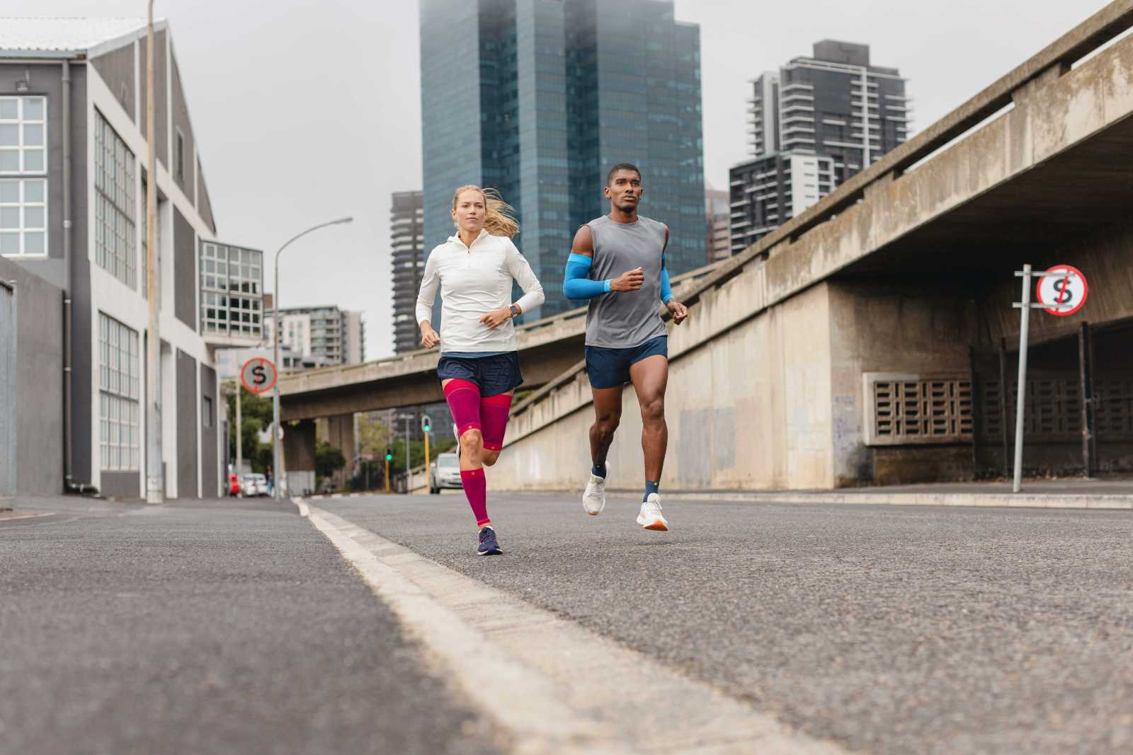 A woman with pink compression sleeves and a man with blue arm sleeves and short running socks run through a big city