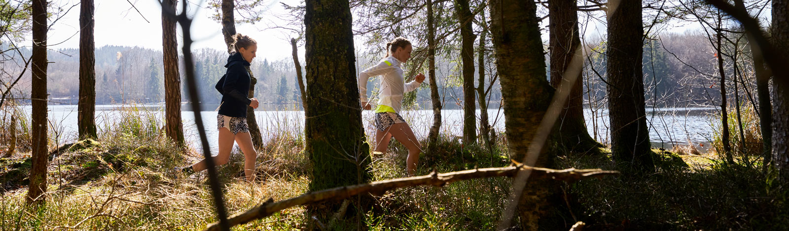 Two runners run behind trees and in front of a small lake in a row over a forest path