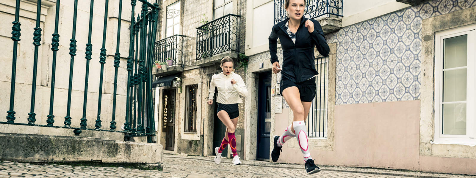 Two women run vigorously through an old town street with small balconies