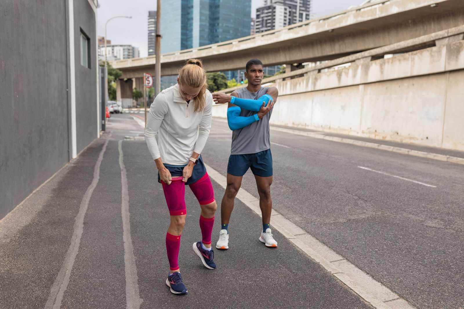 A woman with pink compression Sleeves and a man with blue arm sleeves and short running socks take a break on the roadside of a big city and stretch out