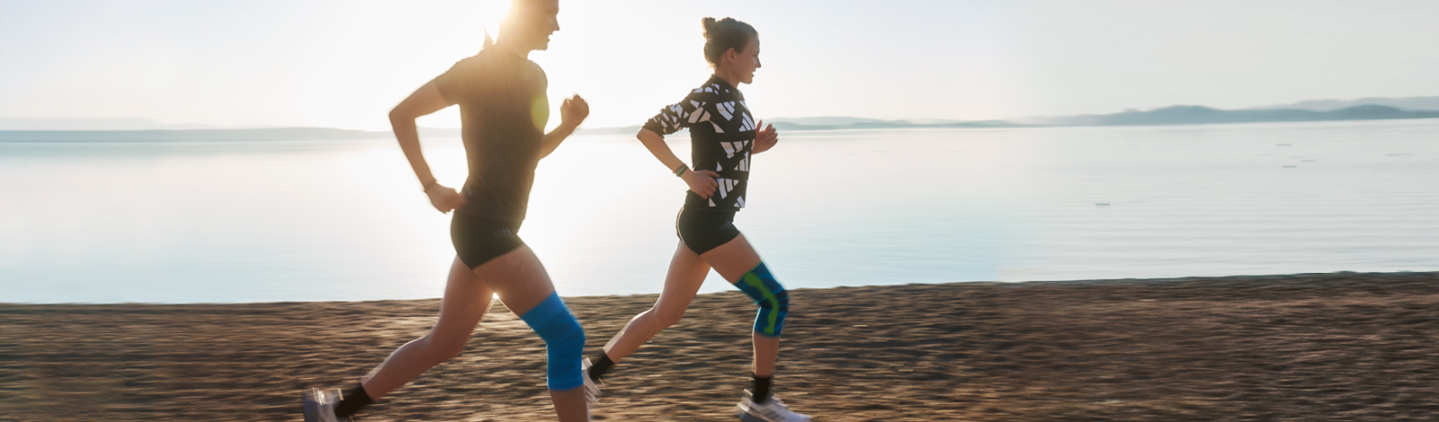 Two runners with knee bands run at a beach next to a lake at dusk