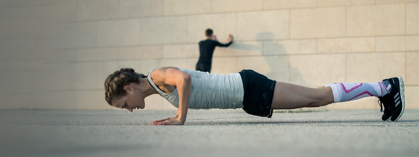 Woman makes push-ups on a concrete floor in the background a man stretches on a wall