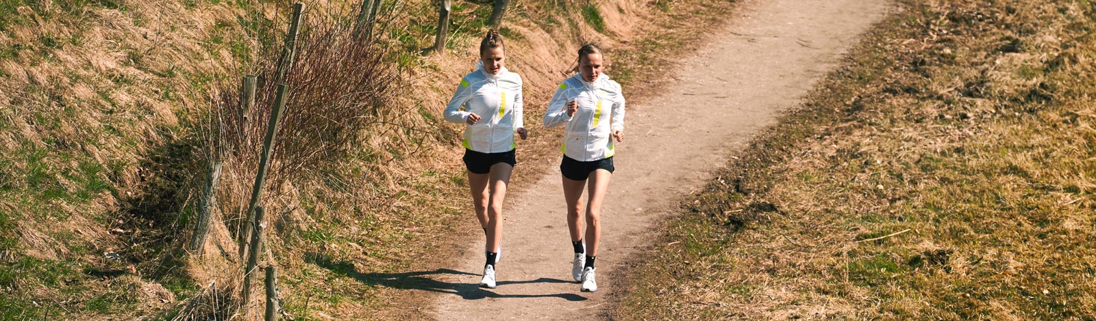 Two runners in bright running jackets run on a narrow path between sparingly overgrown meadows in nature