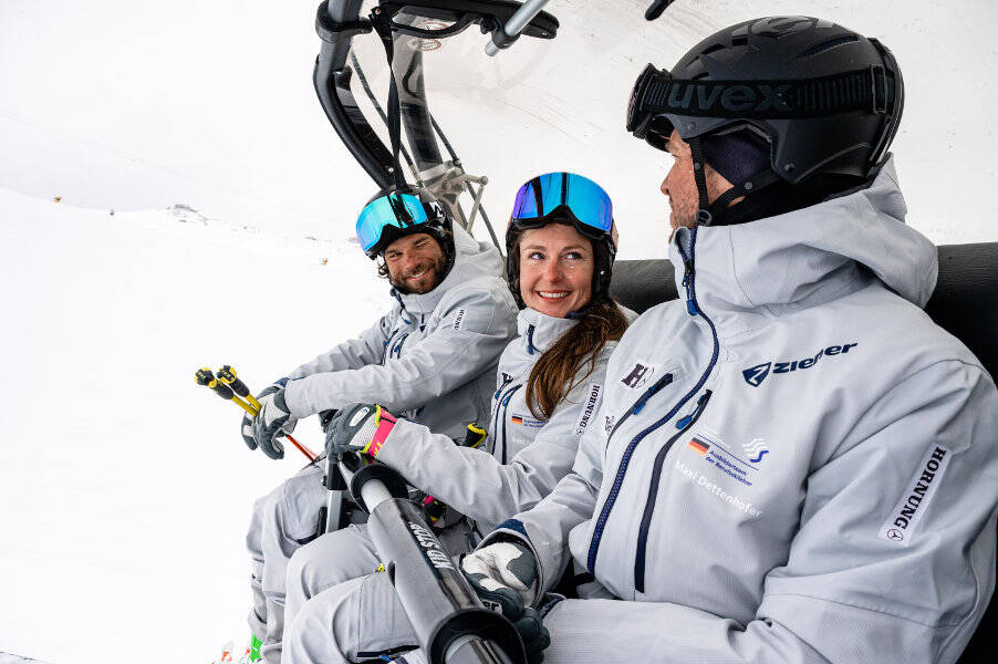 A woman and two men in gray ski suits sit together in a chair lift the woman looks at her colleague with a smile