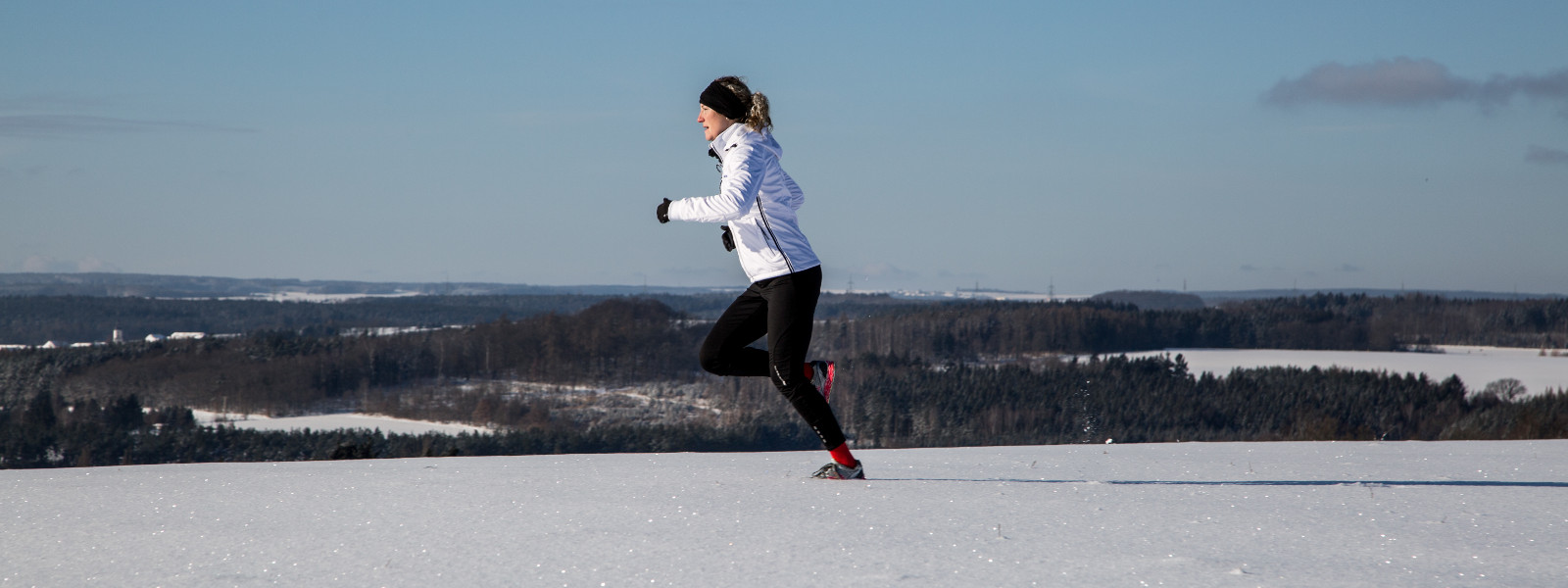 Woman in a white running jacket jogges with even snow field in the background, small forests and clearings alternate