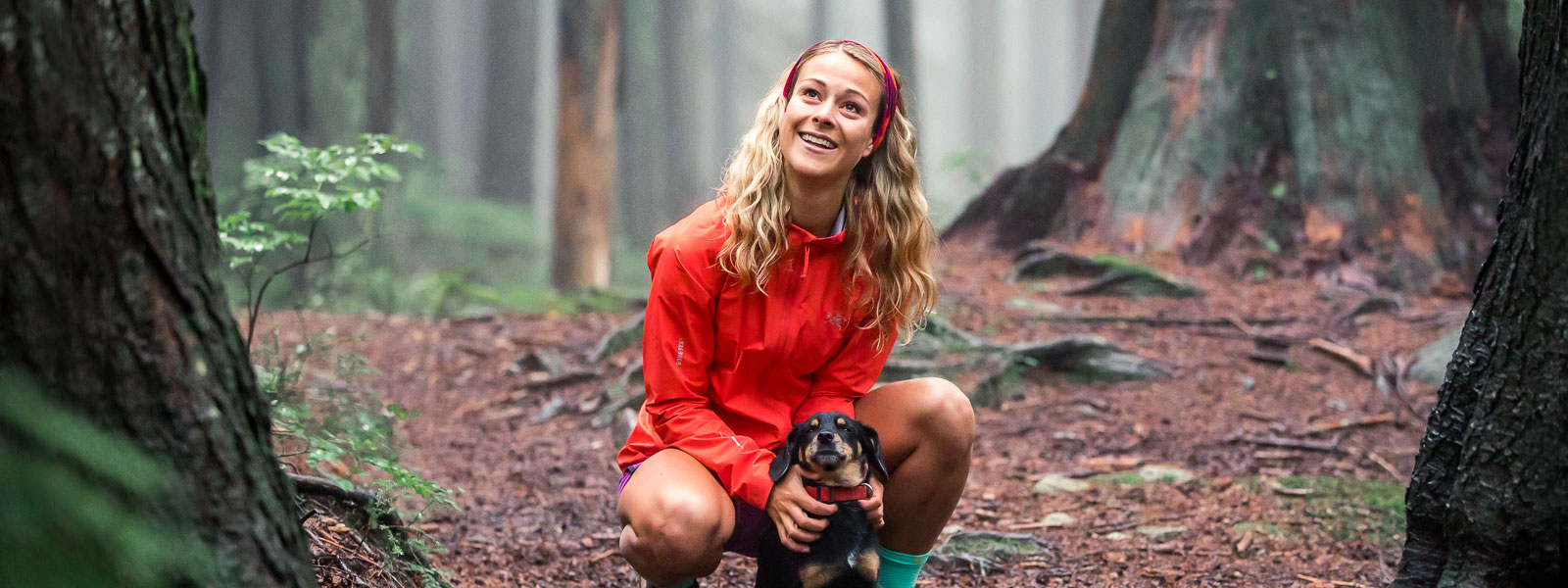 Woman with a red running jacket and headband holds her little dog on a forest path and smiles
