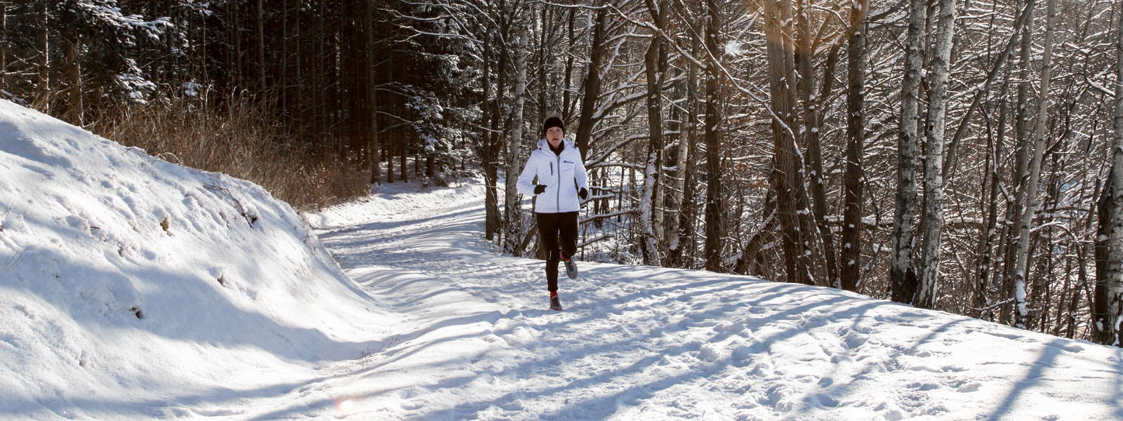 Woman in a white running jacket jogges on a snow -covered path between trees and looks towards the camera
