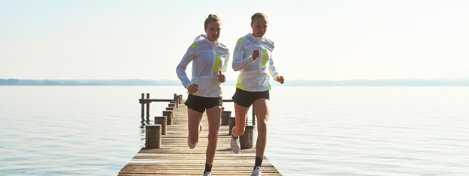 Two runners in white running jackets and with short pants run over a jetty towards the bank