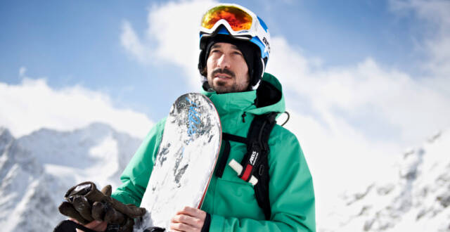 Snowboarder with a green jacket with board in hand in front of a mountain backdrop