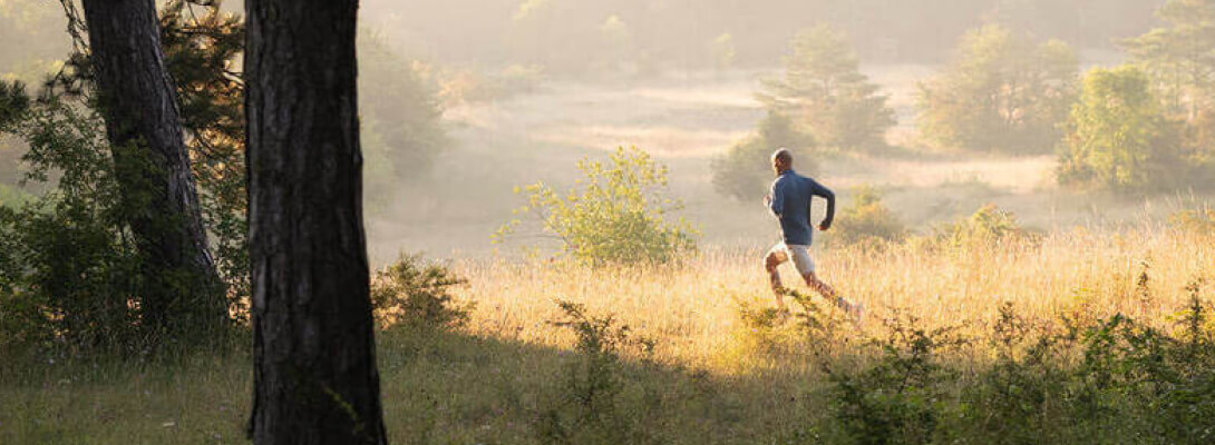 Man jogs over a meadow in the foreground a tree in the background are hills