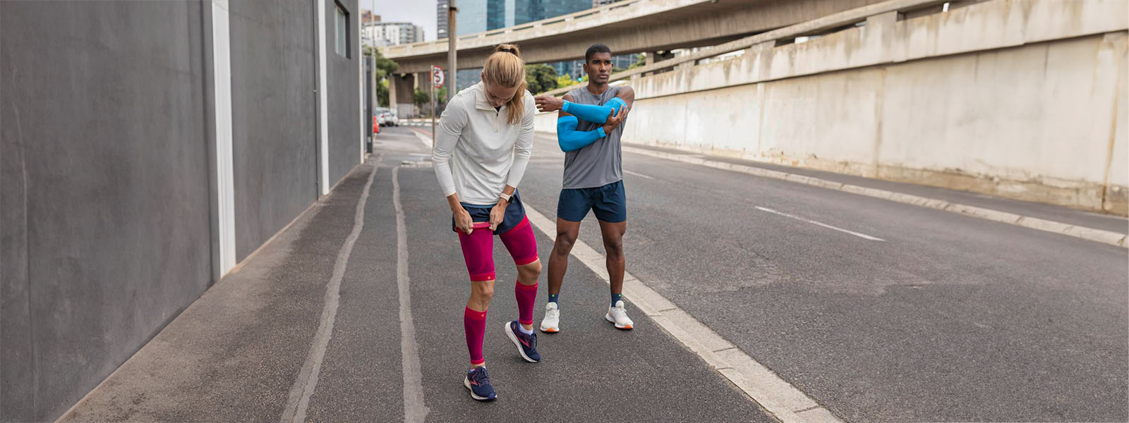 A woman with pink compression Sleeves and a man with blue arm sleeves and short running socks take a break on the roadside of a big city and stretch out