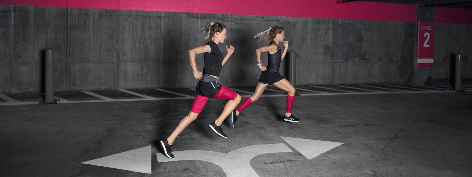 Two runners with pink compression sleeves races through a parking garage