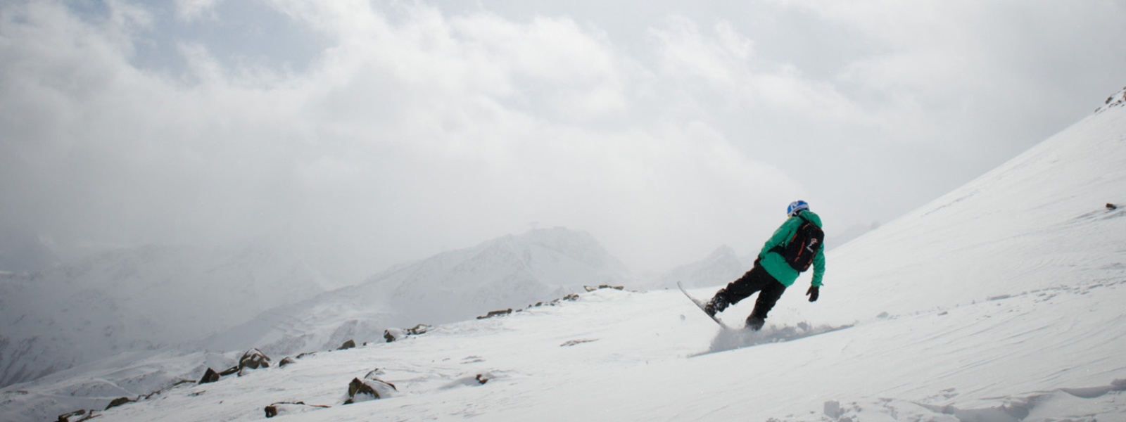 Snowboarders in a green jacket brakes with the rear part of his board in front of a top in the background you can see snowy mountain peaks through the clouds