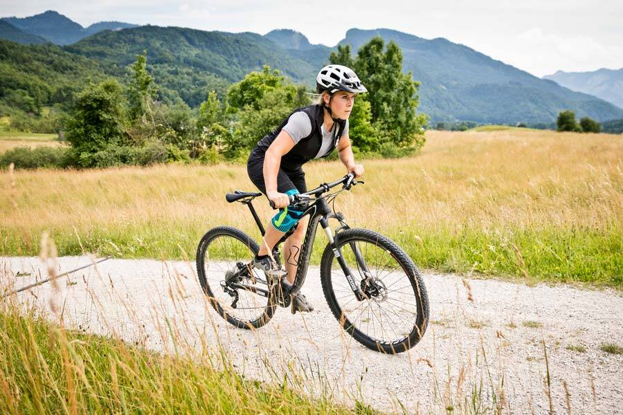 Woman with a kneeboard runs on her mountain bike over a gravel path between meadows and fields