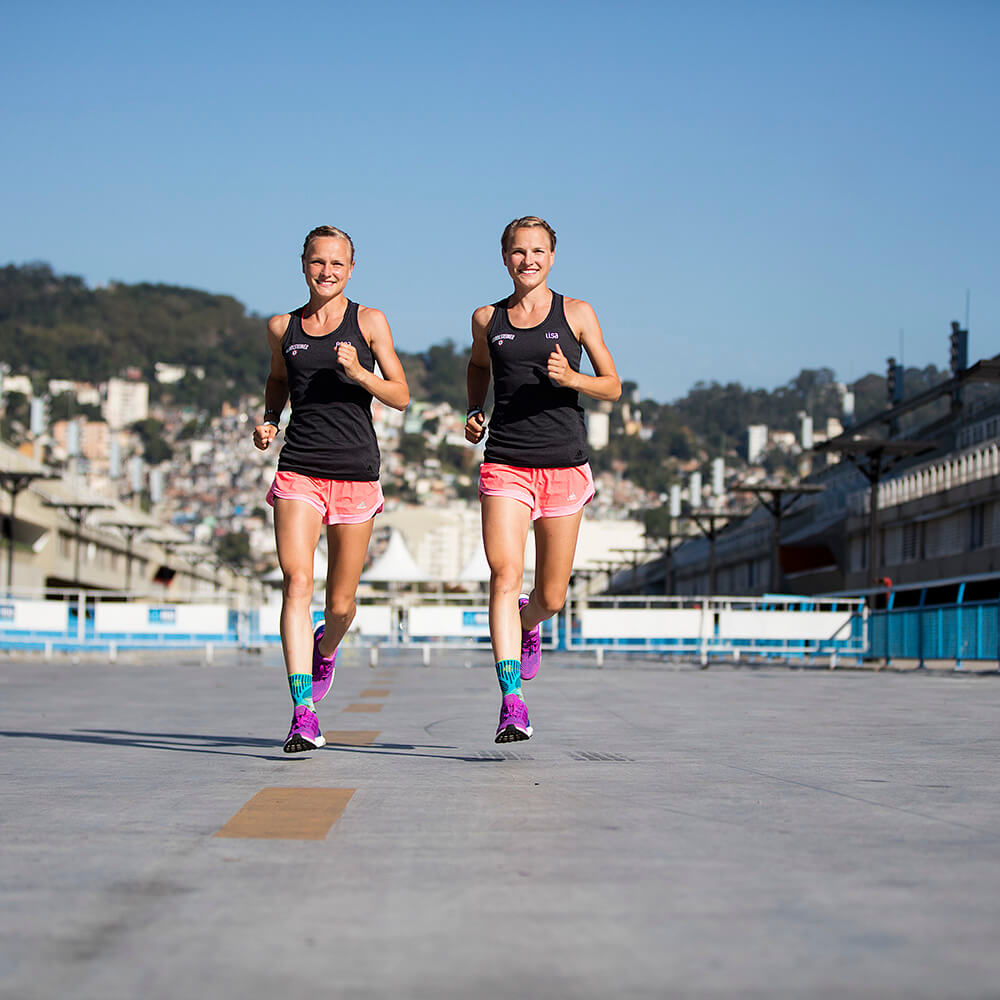 Hahner Twins run on a street with a smile on the camera to carry ankle bandages for sport. A city and hill can be seen in the background.