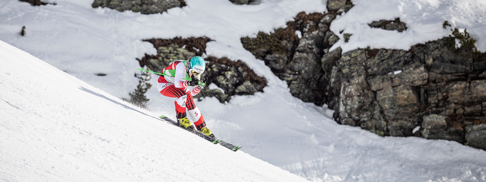 Ski runner in a red and white racing suit in a downhill position with a slight swing with large snow-covered rocks in the background