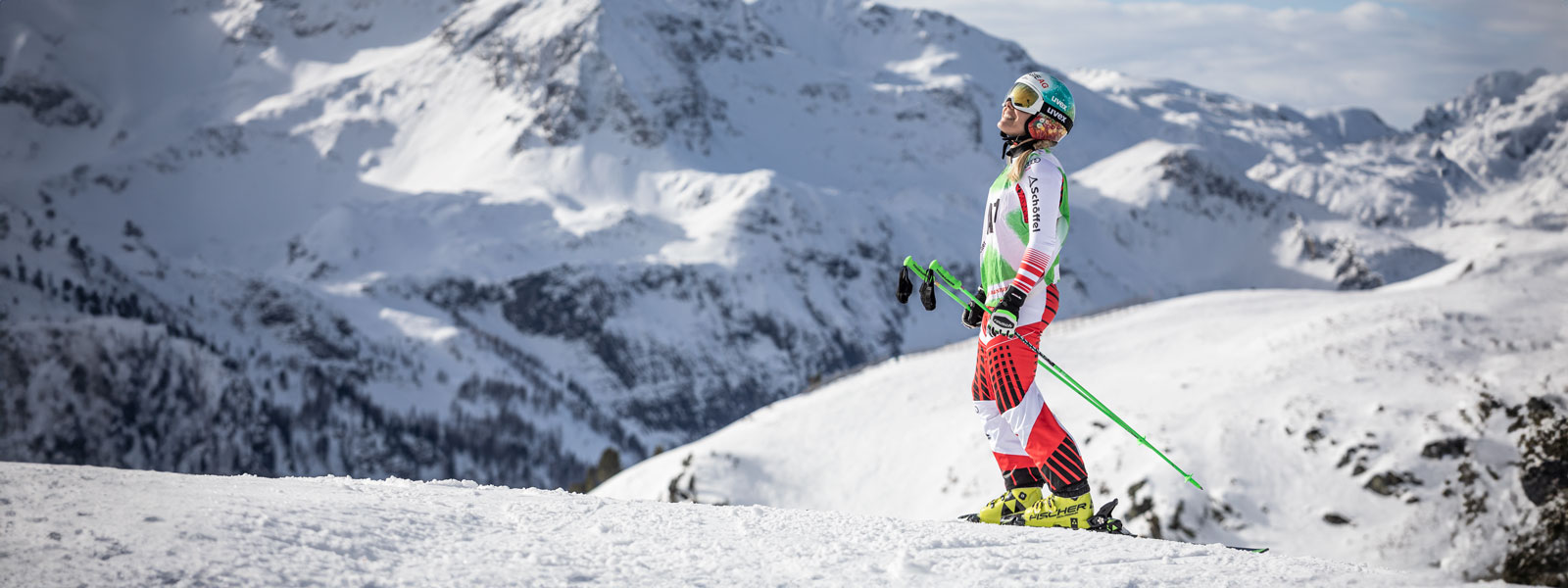 Ski driver in a red and white racing suit is smiling and with the sticks in her hands in front of a huge mountain range