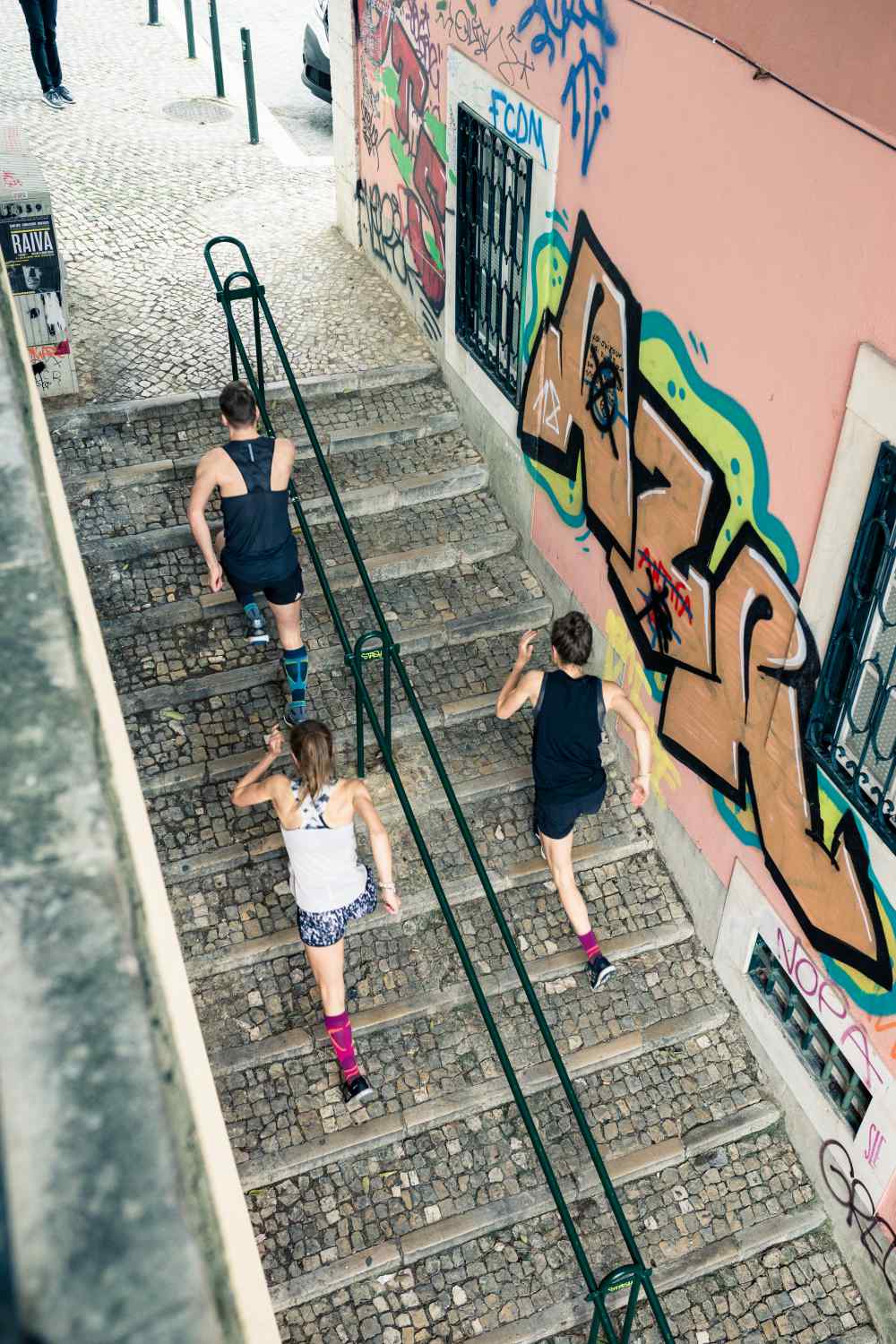 Bird's eye view: A man and two women in running outfits run a staircase in an old town with graffiti on the walls