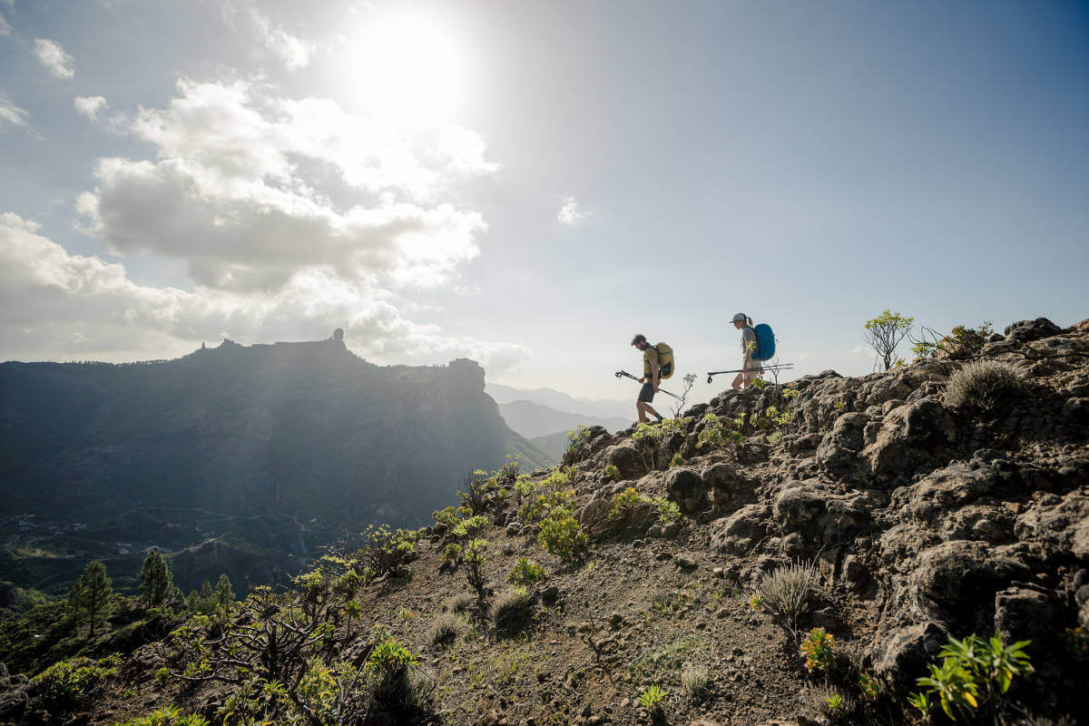 Wide angle: Two hikers with large backpacks and sticks carefully go a mountain range with stones and small shrubs downwards in the background other mountains can be seen
