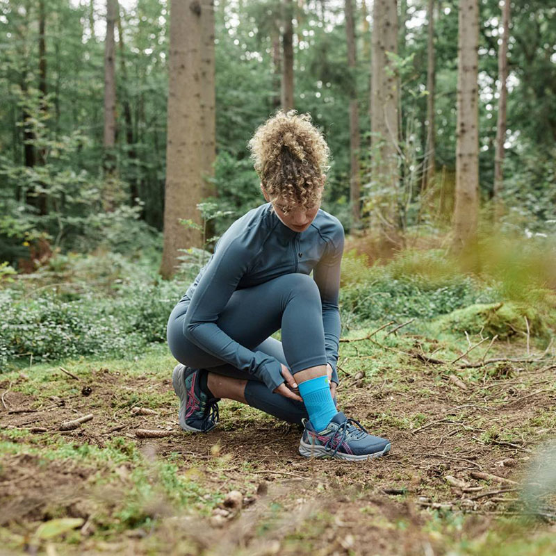 A female runner kneels on the ground in the forest adjusting her Achilles bandage