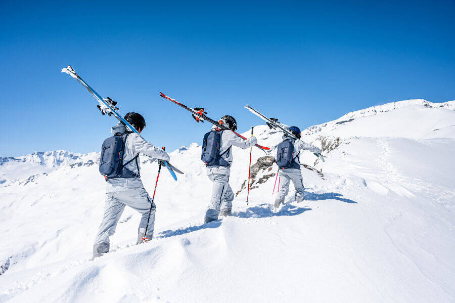 Three skiers carry their skis on their shoulders through deep snow up a mountain